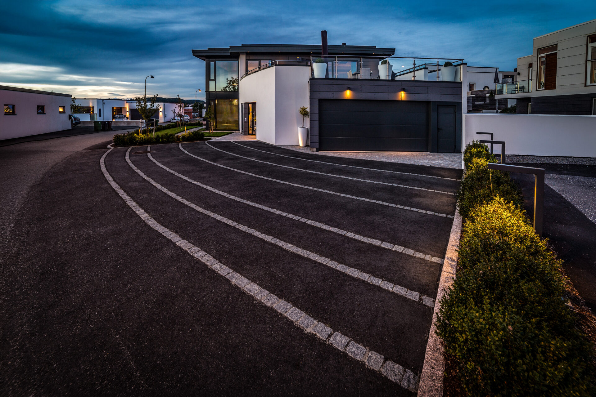 Innkjørsel og adkomst utformet med tydelig linjeføring, der murte kanter og faste overflater binder bolig, garasje og utearealer sammen i et helhetlig uttrykk